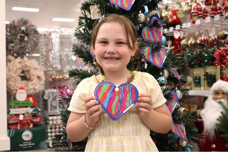 A young girl with light brown hair grins at the camera, holding a colourful heart that she designed. She wears a yellow dress, has her hair in plaits and stands in front of a Christmas tree.